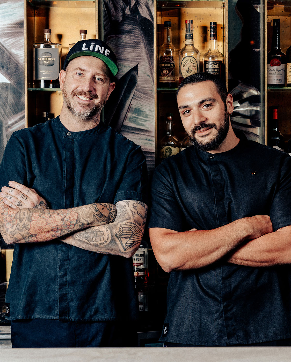 Two men stand side by side in front of shelves filled with various bottles. Both are wearing dark chef coats. The man on the left, with tattoos and a baseball cap, has his arms crossed. The man on the right, with a beard, also crosses his arms and smiles.