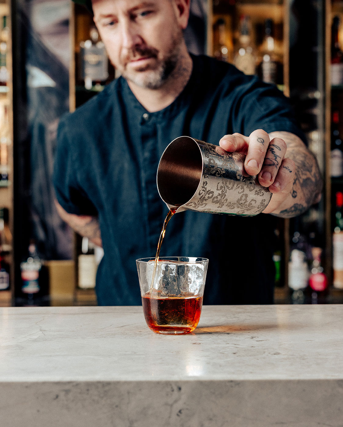 A person with tattoos and a beard pours a beverage from a metal shaker into a glass at a bar. The countertop is marble, and shelves with various bottles are in the background.