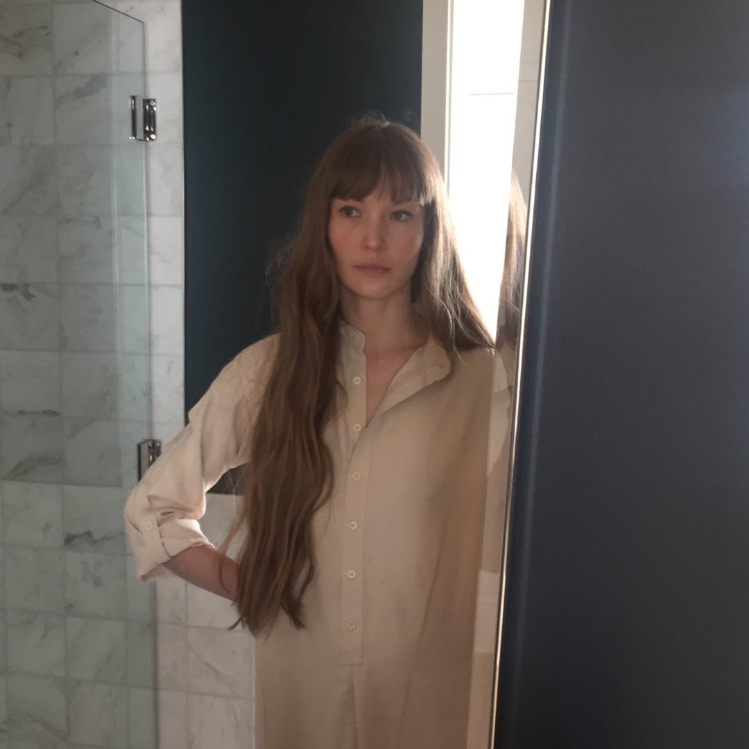 A woman with long, straight hair and bangs stands in front of a bathroom mirror, wearing a light-colored, long-sleeve button-up shirt. She looks off to the side with a neutral expression. The bathroom features marble tiled walls and a glass shower enclosure.