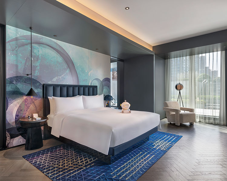 Chambre d'hôtel moderne avec un grand lit, une tête de lit bleue et des motifs géométriques sur le mur. Un tapis à motifs bleus repose sur le plancher en bois. Il y a un fauteuil de couleur claire près de la fenêtre avec vue sur la ville et une petite table d'appoint avec une lampe à côté du lit.