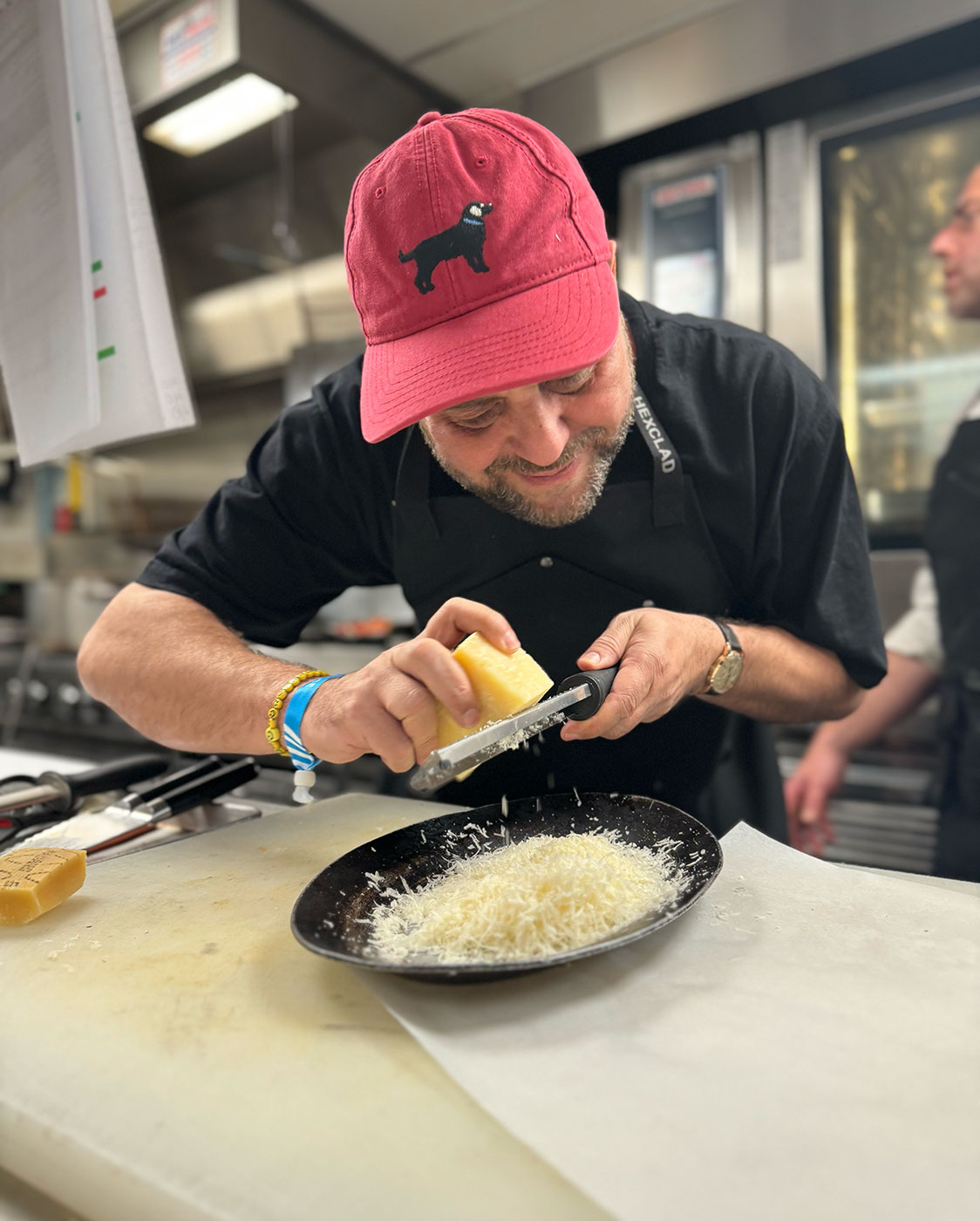 A man, wearing a red cap and a black apron, grates cheese over a black plate. He is focused on his task in an industrial-style kitchen. The background shows another person partially visible and kitchen equipment.