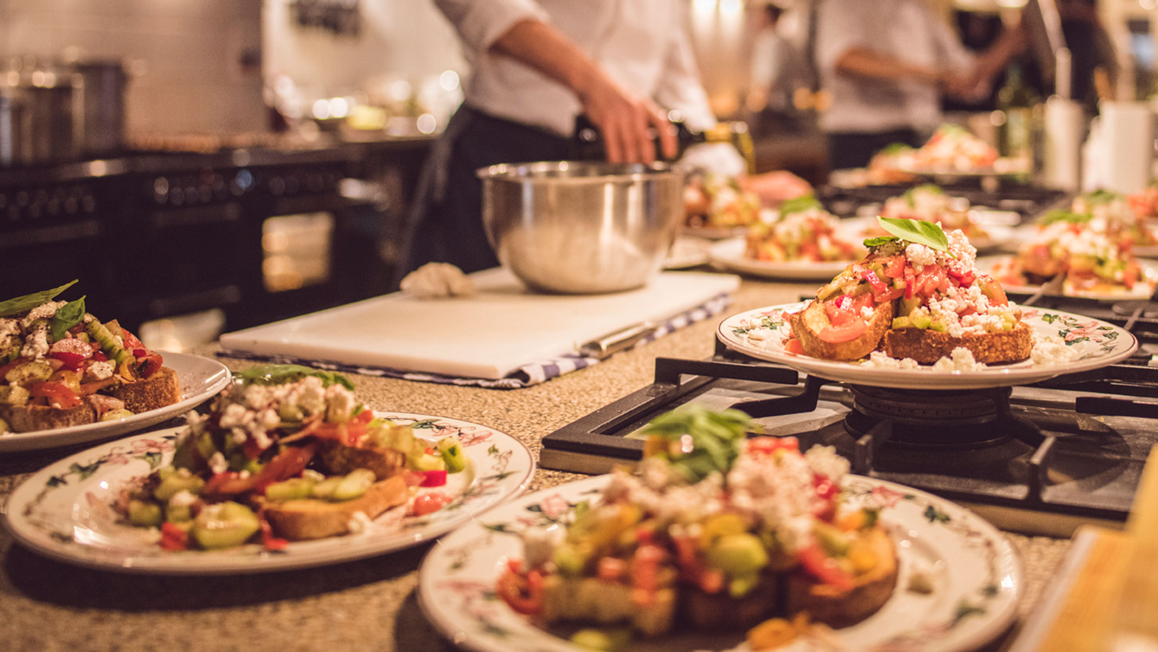 A chef prepares multiple plates of colorful, fresh salads in a professional kitchen. Each plate is full of vibrant vegetables, topped with crumbled cheese and garnished with basil. The kitchen environment is bustling, with other chefs working in the background.