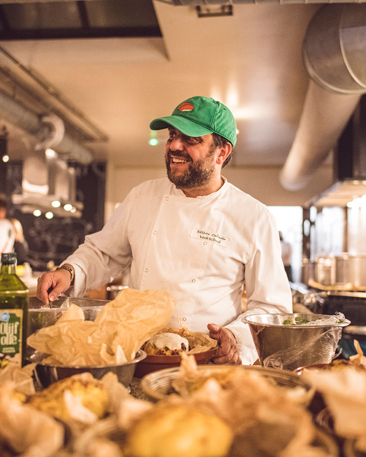 A smiling chef in a white uniform and green baseball cap stands in a busy kitchen, preparing food. Surrounding him are various ingredients and cooking utensils. The atmosphere appears lively and bustling.