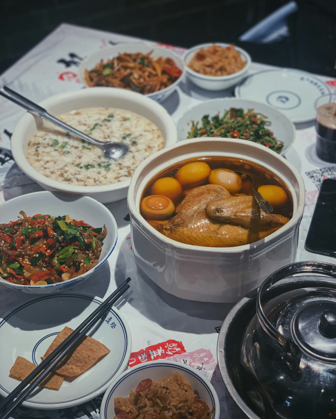 A table filled with various Chinese dishes, including a whole chicken in broth, a pot of rice congee, several bowls of stir-fried vegetables, pickled vegetables, and tofu. Chopsticks, a spoon, and plates are also arranged on the table.