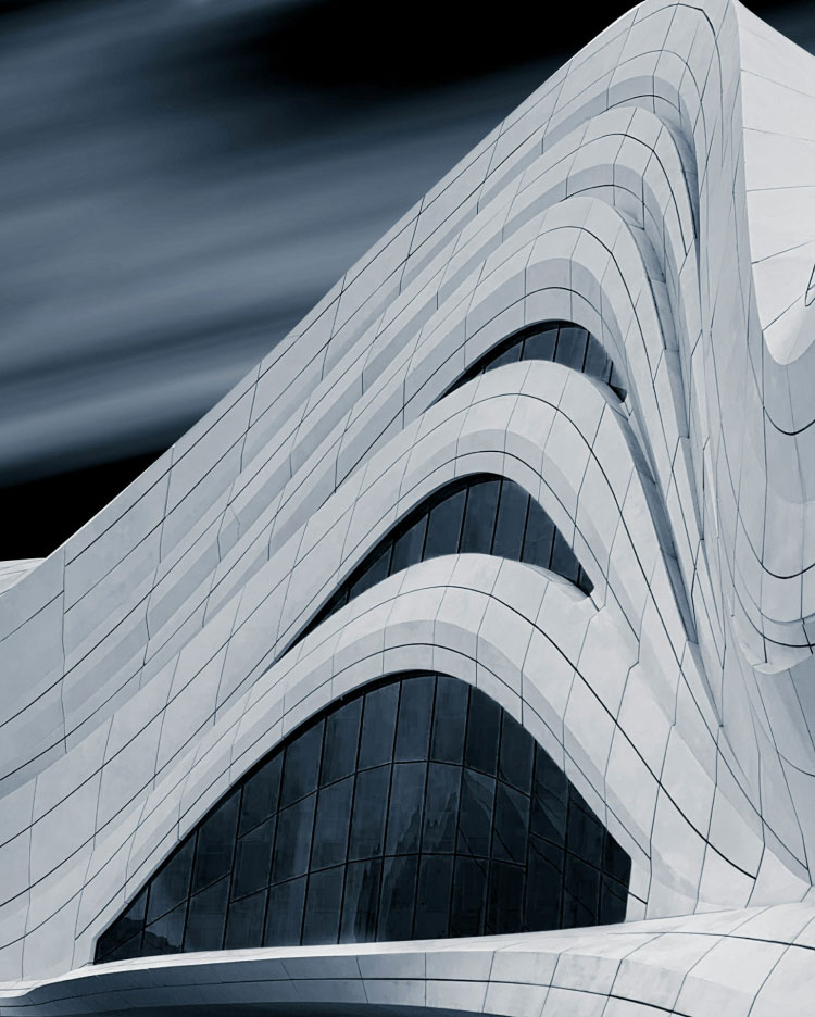 A modern, abstract building with curving, asymmetrical lines and large, glossy windows. The facade features a series of smooth, white, wave-like ridges that give it a futuristic appearance. The sky is a blurred mix of light and dark hues, enhancing the dramatic effect.