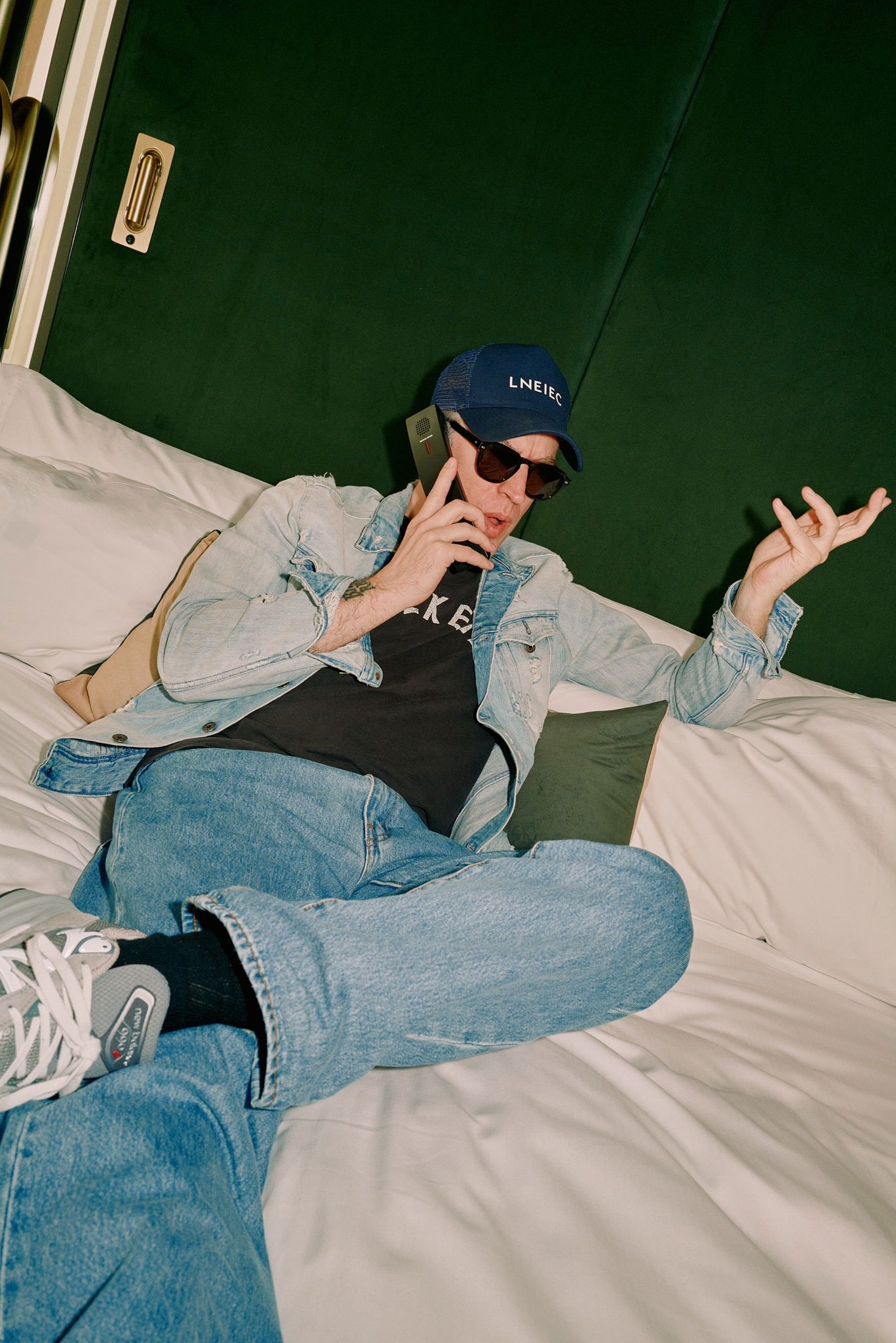 A person in a denim jacket, jeans, sneakers, and a dark cap and sunglasses lounges on a bed while talking on a smartphone. They appear relaxed, holding the phone to their ear with one hand and gesturing with the other. The background is a dark-colored wall.