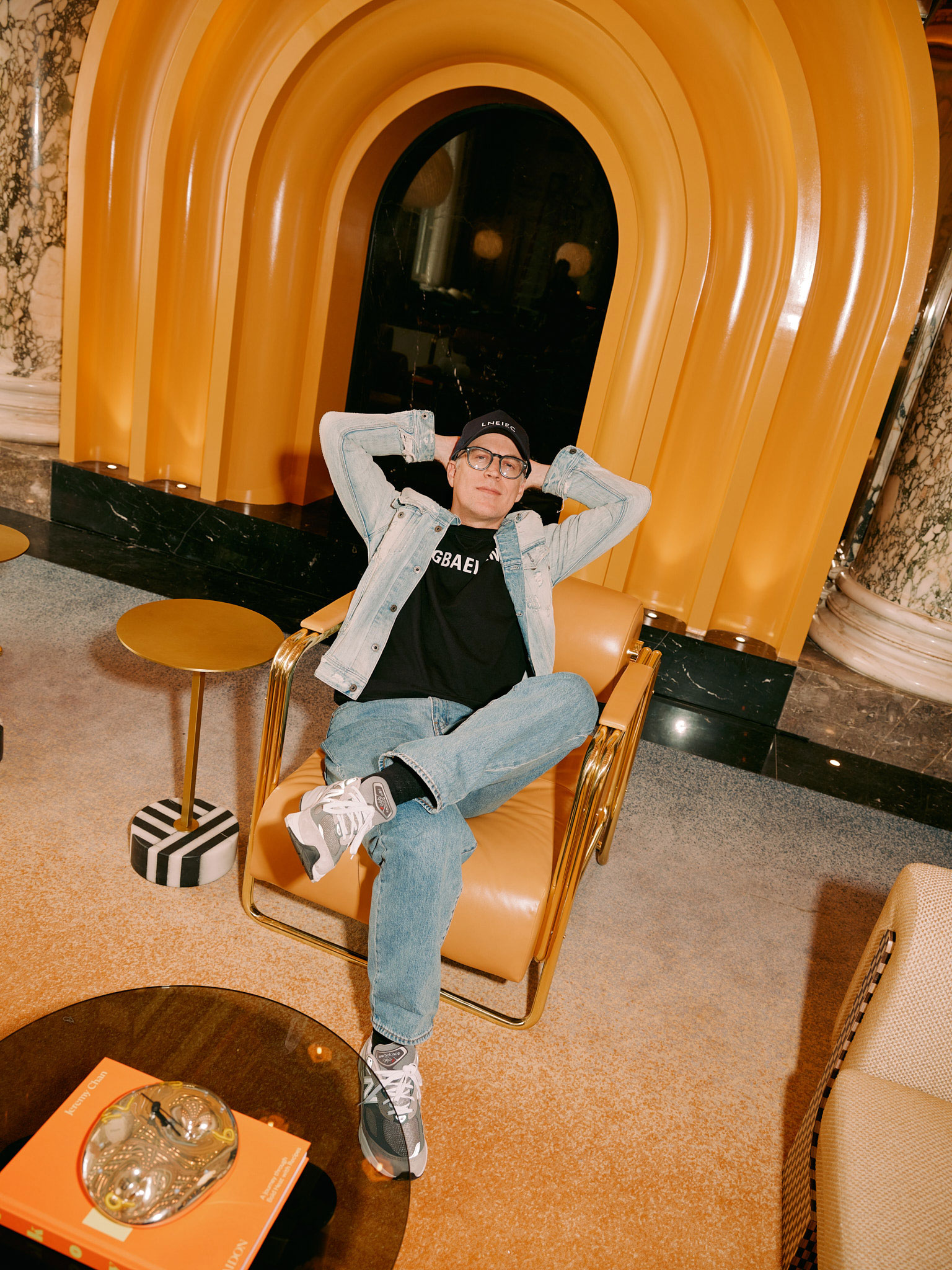 A person sits relaxed in a stylish chair in front of an archway with orange accents. They wear a cap, glasses, a black shirt, and denim jacket and jeans. The room features a table with books and decorative items, and marble columns on the sides.