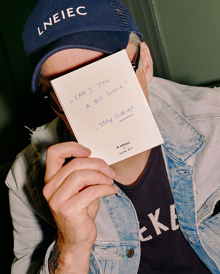 A person wearing a cap and sunglasses holds a note in front of their face. The note reads, 