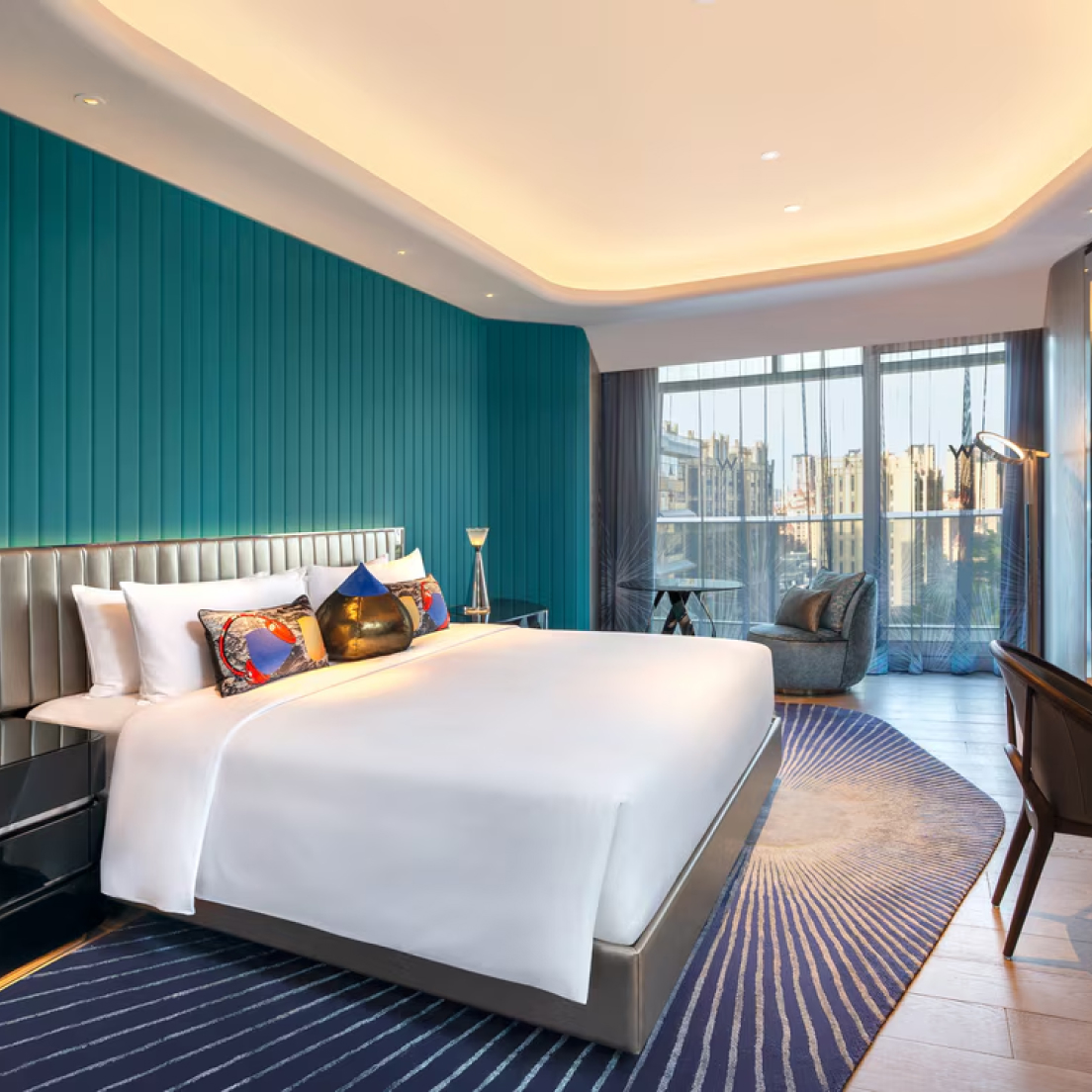 Habitación de hotel moderna con una cama grande con sábanas blancas y almohadas de colores. Pared con detalles en azul, ventana grande con vista a la ciudad, sillón gris y escritorio de madera con silla. La suave iluminación del techo crea un ambiente acogedor.