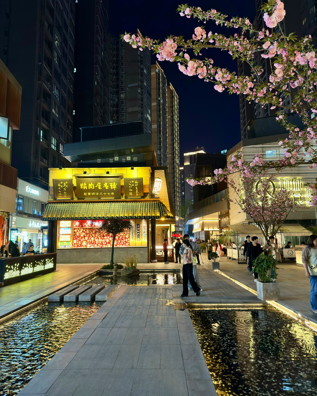 A bustling, well-lit urban street at night features traditional-style buildings with illuminated signs. Cherry blossoms on a nearby tree add a touch of nature. Pedestrians walk along the path, bordered by shallow rectangular pools of water reflecting the city lights.