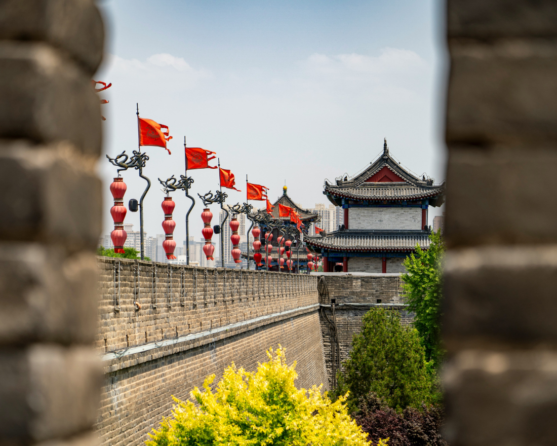 View through a gap in a stone wall of a historic building with traditional Chinese architecture. Red lanterns and flags adorn the wall, and lush green trees are visible in the foreground under a clear blue sky.