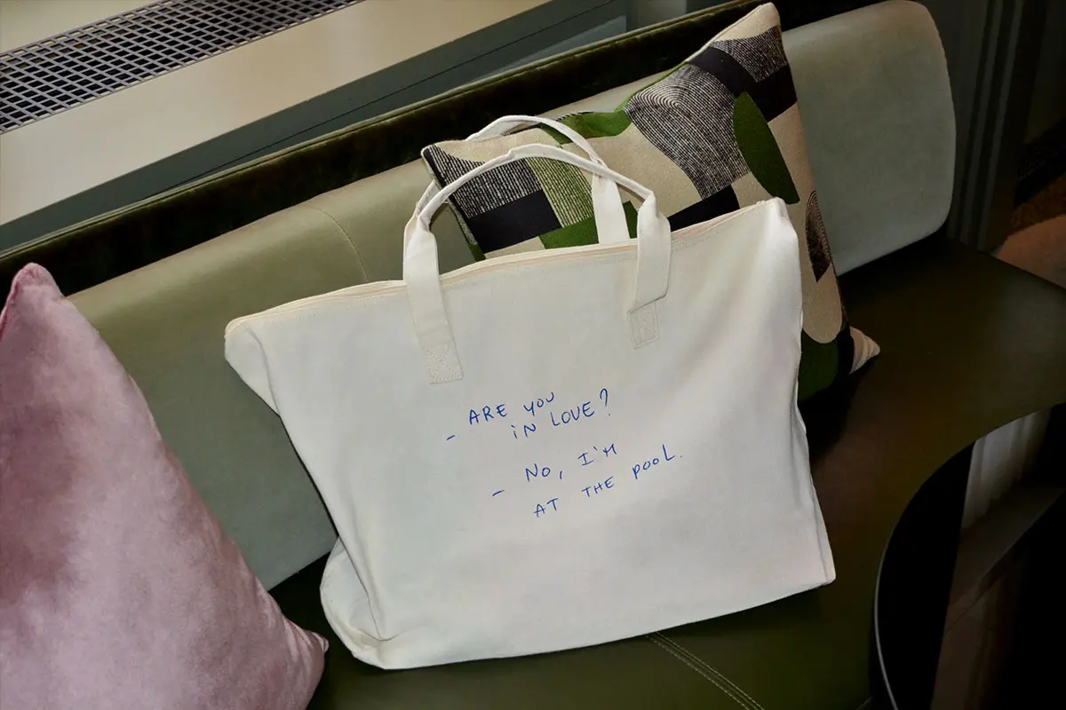 A white tote bag resting on a green cushioned bench with a purple pillow and patterned pillow beside it. The bag has blue handwritten text that reads: 