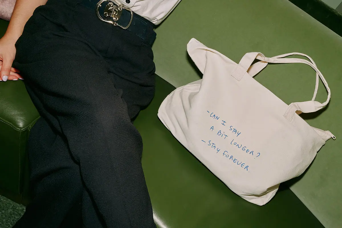 A person wearing black pants and a white shirt is sitting on a green couch. Next to them is a white tote bag with text that reads, 