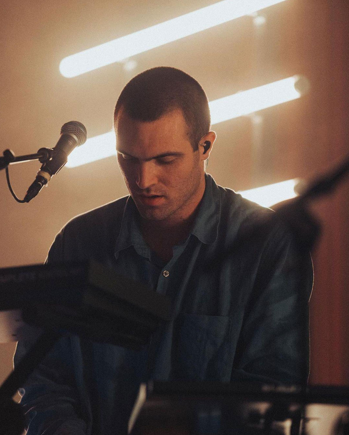 A person in a dimly lit room plays a keyboard with a microphone nearby. They are wearing a button-up shirt, and there are vertical lights behind them creating a warm atmosphere.