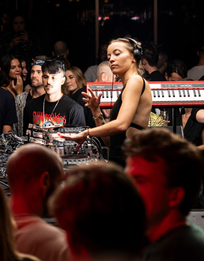 A DJ wearing headphones is performing in a dimly lit club. She is surrounded by music equipment, including keyboards and a mixer. A crowd of people is gathered around, some with drinks in hand, enjoying the music.