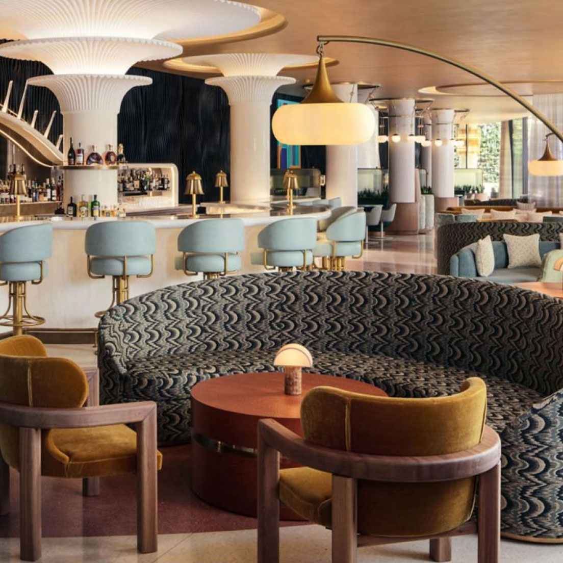 Eine stilvolle Hotellobbybar mit einem geschwungenen gemusterten Sofa, runden Tischen mit Lampen und senfgelben Stühlen. Der Barbereich verfügt über hellblaue gepolsterte Hocker und ein elegantes, modernes Design mit einer gewölbten goldenen Leuchte.