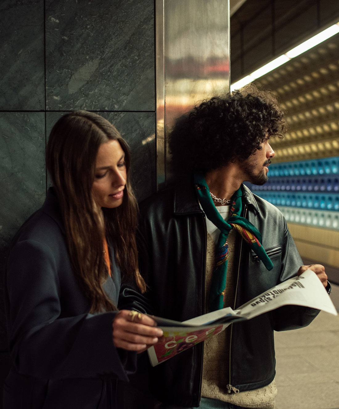 Una mujer y un hombre están de pie en una estación de metro. La mujer está mirando un periódico y el hombre está de espaldas, mirando hacia el andén. Ambos llevan chaquetas y el andén tiene un diseño moderno con hileras de luces.