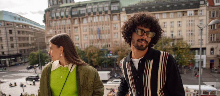 A man and a woman stand outdoors in an urban setting with historic buildings in the background. The woman, wearing a green top and olive jacket, looks to the side, while the man, in sunglasses and a striped shirt, faces forward.