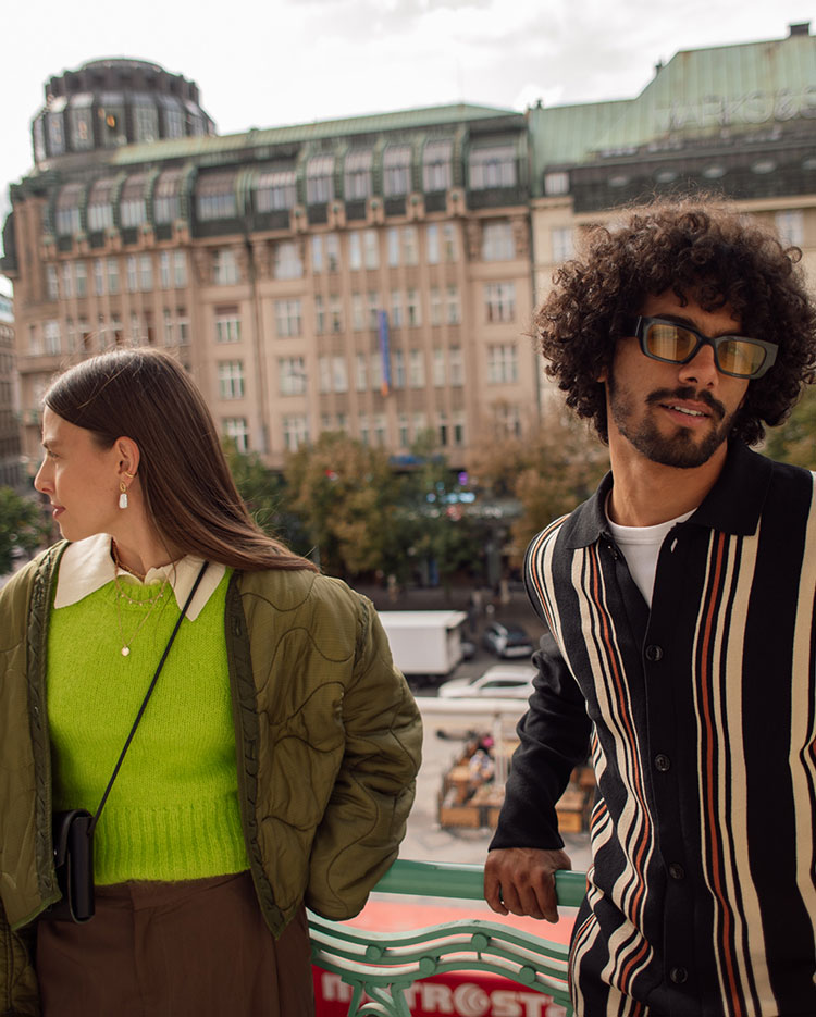 A man and woman stand on a balcony overlooking a city street. The woman wears a green sweater and quilted jacket, glancing sideways. The man wears sunglasses and a striped shirt, leaning on the railing. A historic building is in the background.