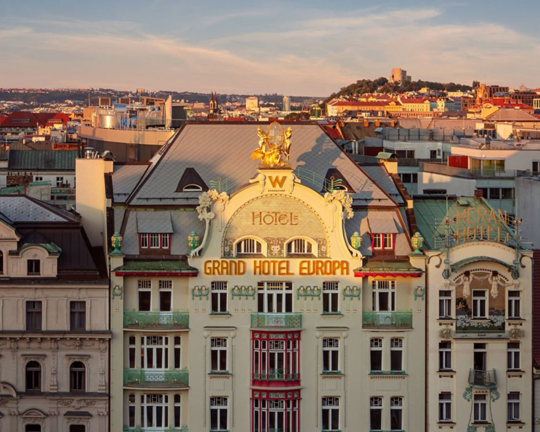 A view of a European cityscape featuring the Grand Hotel Europa with its ornate architecture and golden statue on the roof. Buildings surround the hotel, and a hill with a structure on top is visible in the distant background under a clear sky.