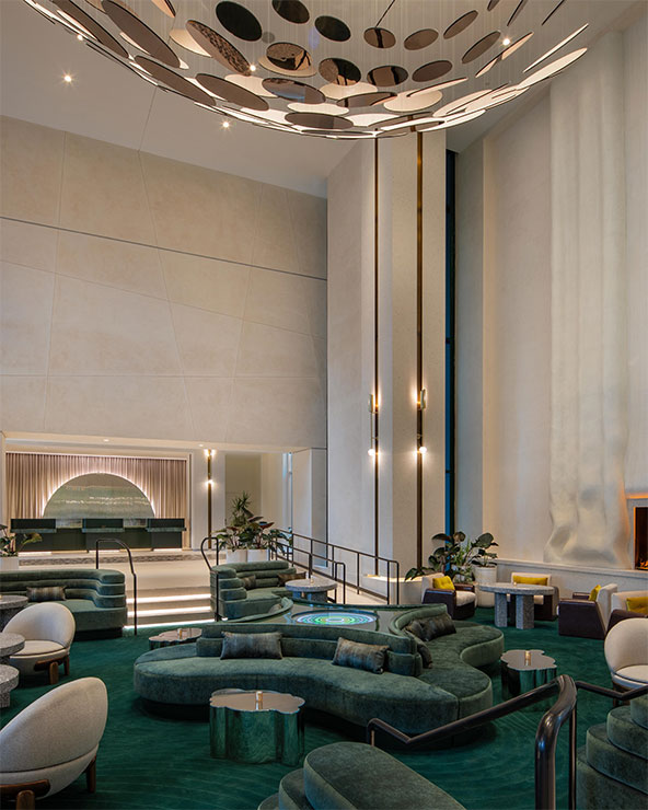 A modern hotel lobby featuring curved green velvet seating, circular tables, and a dramatic ceiling installation with reflective circles. Tall cream walls and vertical light fixtures add to the elegant ambiance.