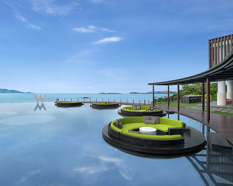 A luxurious outdoor seating area with round cushioned lounges on a reflective water surface. The scene overlooks a tranquil sea and a clear blue sky. A wooden deck extends towards the horizon, offering a serene coastal view.