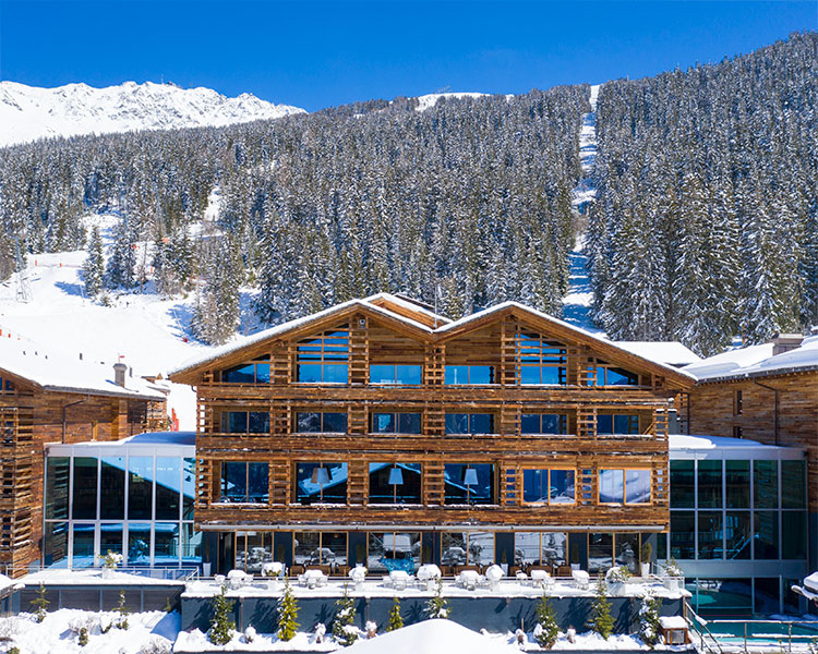 A large wooden lodge with numerous windows is set against a backdrop of snow-covered pine trees and a mountain. The sky is clear and blue, and there's snow on the ground surrounding the lodge.