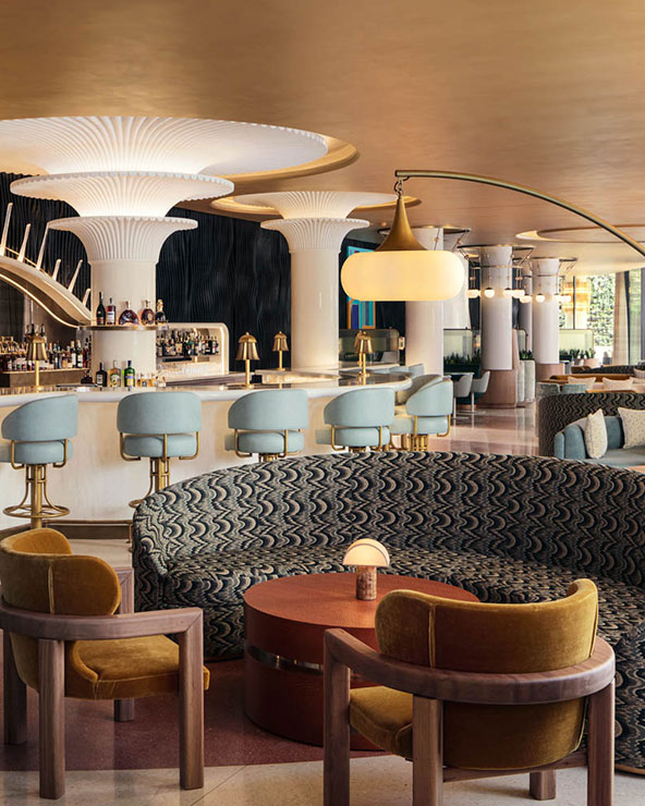 A stylish bar interior with a modern, artistic design. Features include a curving bar with blue stools, unique pillar-shaped structures, retro-patterned benches, and elegant round tables. Warm lighting enhances the inviting atmosphere.