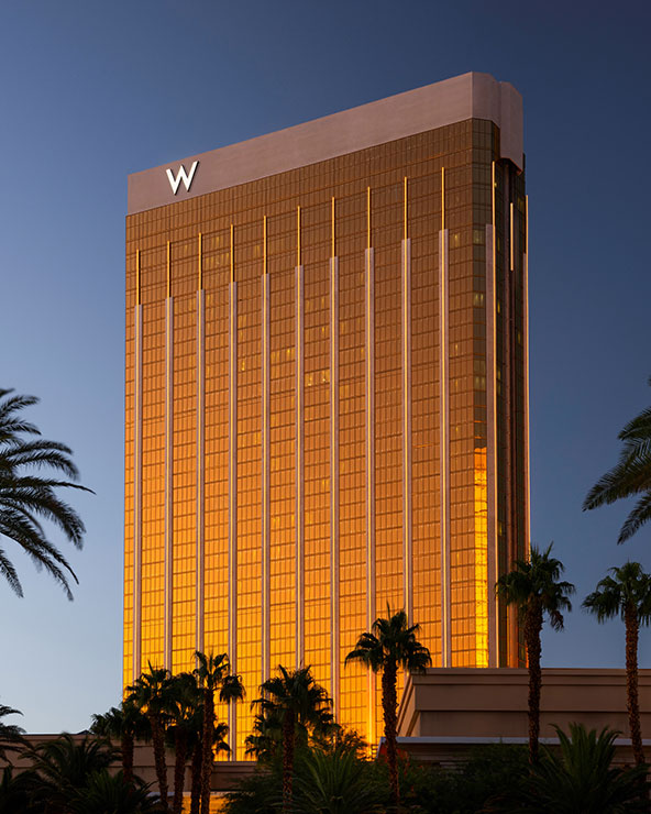 Ein hohes, goldfarbenes Hotelgebäude reflektiert das Abendlicht. An der Spitze ist ein markantes „W“-Logo zu sehen. Im Vordergrund sind Palmen zu sehen und der Himmel wirkt düster.