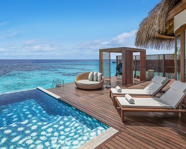 A luxurious oceanfront deck featuring two lounge chairs, a round daybed, and a private infinity pool overlooking the clear blue sea. A thatched cabana and wooden accents add to the tropical ambiance under a bright sky.