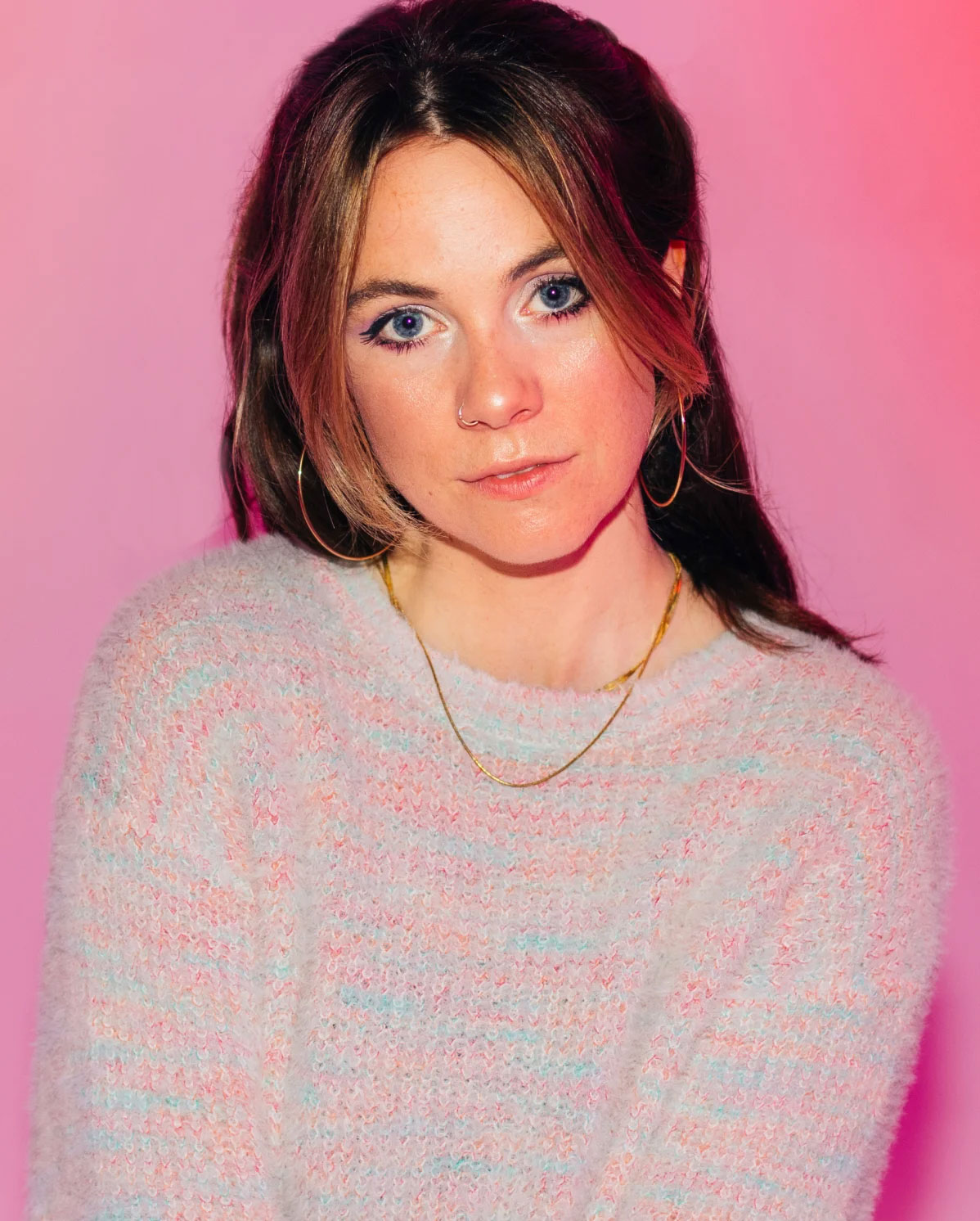 Woman with long brown hair and hoop earrings wearing a fuzzy pastel sweater and a gold necklace. She stands against a pink background, looking at the camera with a slight smile.