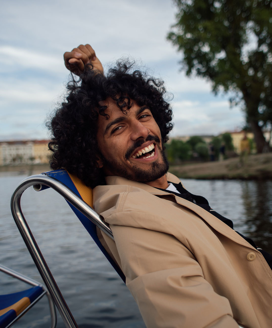 Un hombre de pelo rizado y chaqueta beige sonríe recostado en una silla junto al agua, con árboles y edificios de fondo.