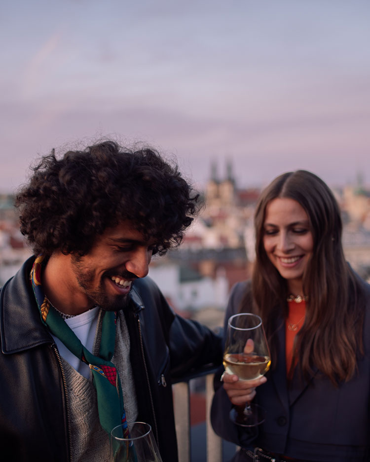 Dos personas sonríen y sostienen copas de vino blanco, de pie, muy juntas, en una azotea al atardecer, con el horizonte de la ciudad difuminado al fondo.