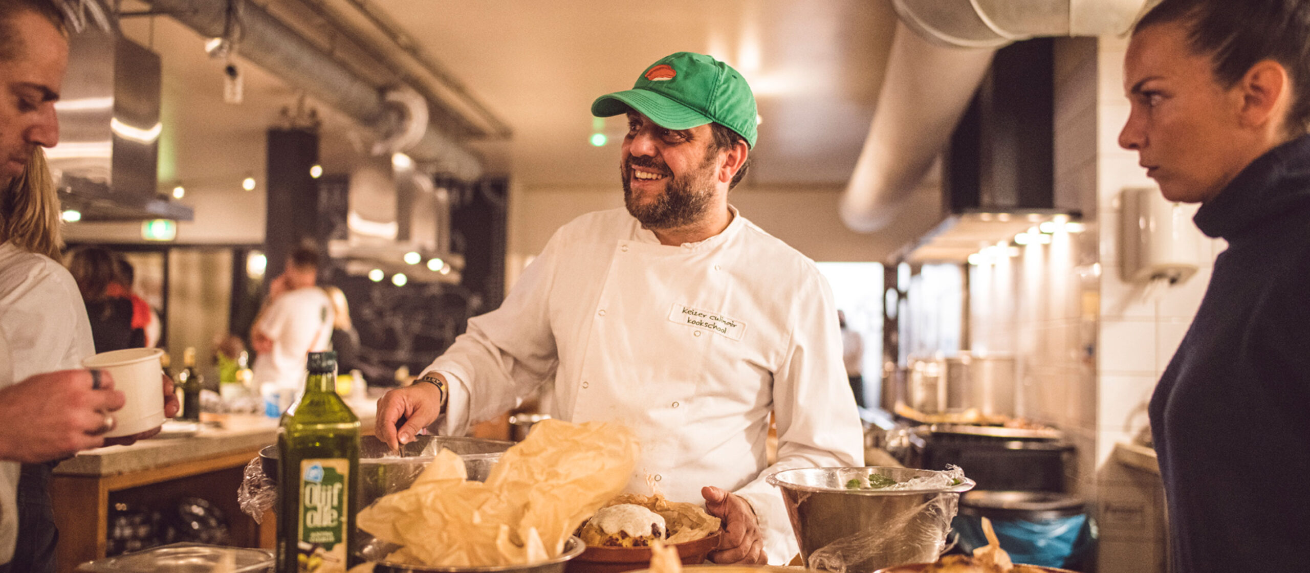 Un chef en tenue blanche et casquette verte sourit tout en préparant de la nourriture dans une cuisine animée. Divers ingrédients et ustensiles de cuisine sont visibles sur le comptoir. Trois autres personnes sont présentes, l'une tenant un bol et l'autre portant un sweat à capuche sombre.