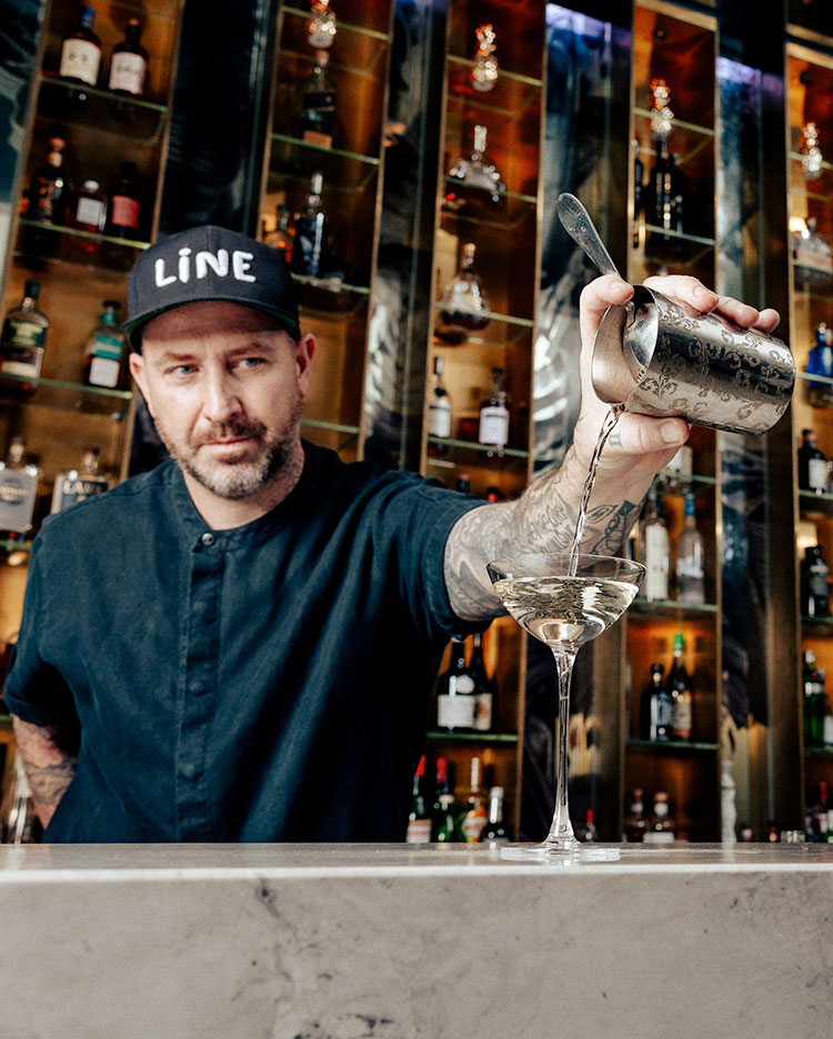 Barman avec une barbe et une casquette LINE versant un liquide clair dans un verre à cocktail à partir d'un shaker dans un bar élégant, avec des bouteilles visibles sur des étagères en arrière-plan.