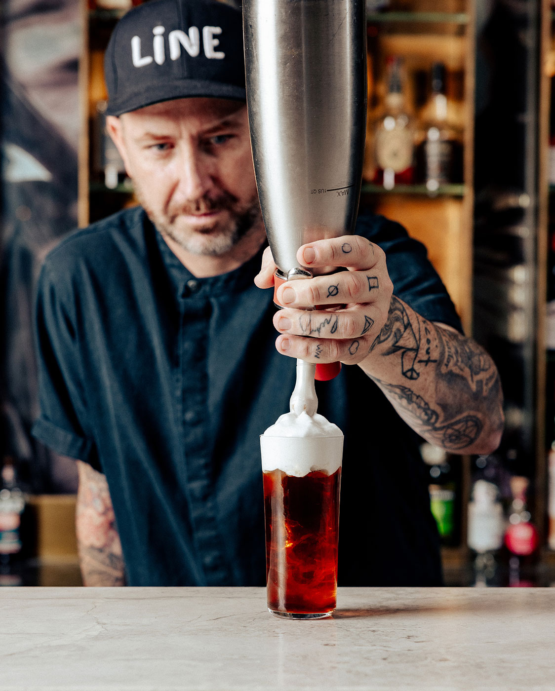 Une personne tatouée, portant une casquette Line et une chemise sombre, verse de la crème fouettée dans un grand verre de boisson sombre dans un décor de bar. Des bouteilles sont visibles dans l'arrière-plan flou.