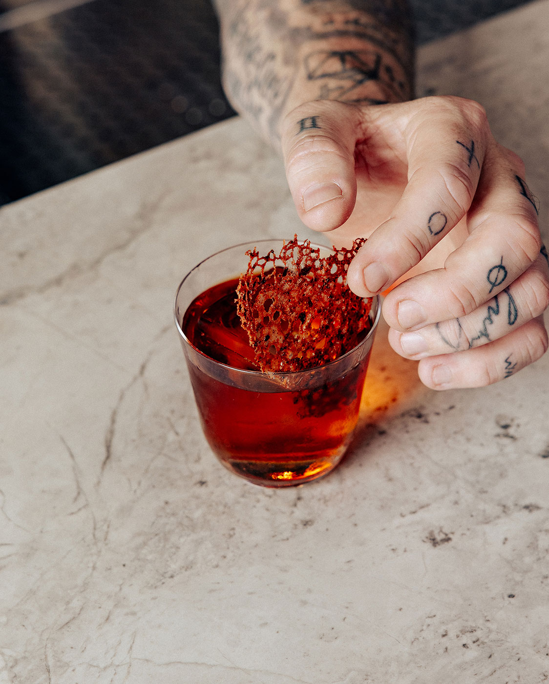 Une main tatouée tient un verre de liquide rouge sur lequel repose une garniture décorative. La boisson est posée sur un comptoir en marbre.