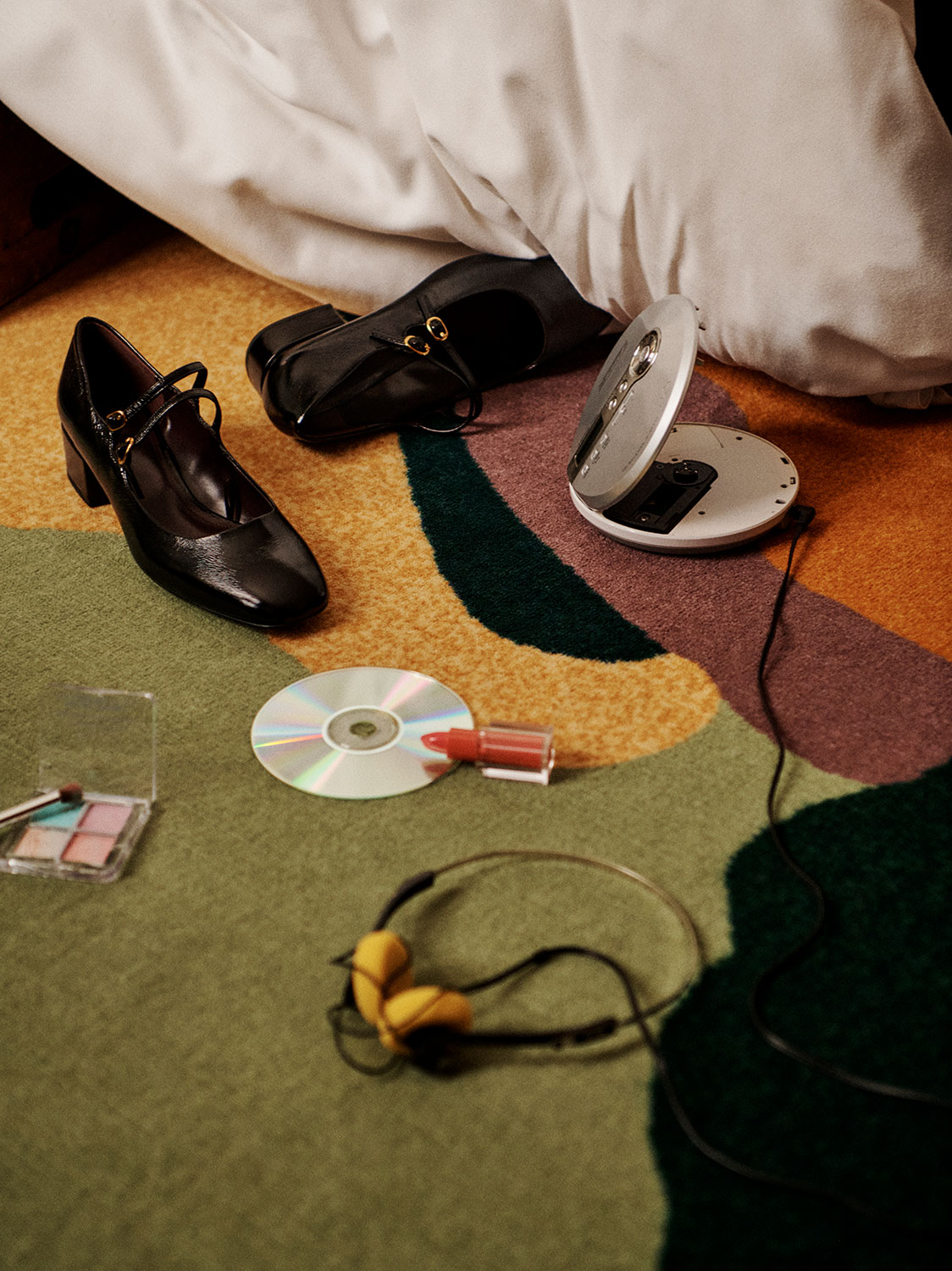 A colorful carpet with a pair of black heels, a portable CD player, headphones, a CD, eyeshadow, and lipstick scattered on it. A white sheet partially covers one shoe.