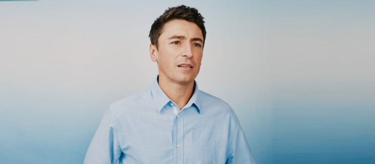 A man with short brown hair wearing a light blue button-up shirt stands in front of a blue and white gradient background, looking slightly to the side with a neutral expression.