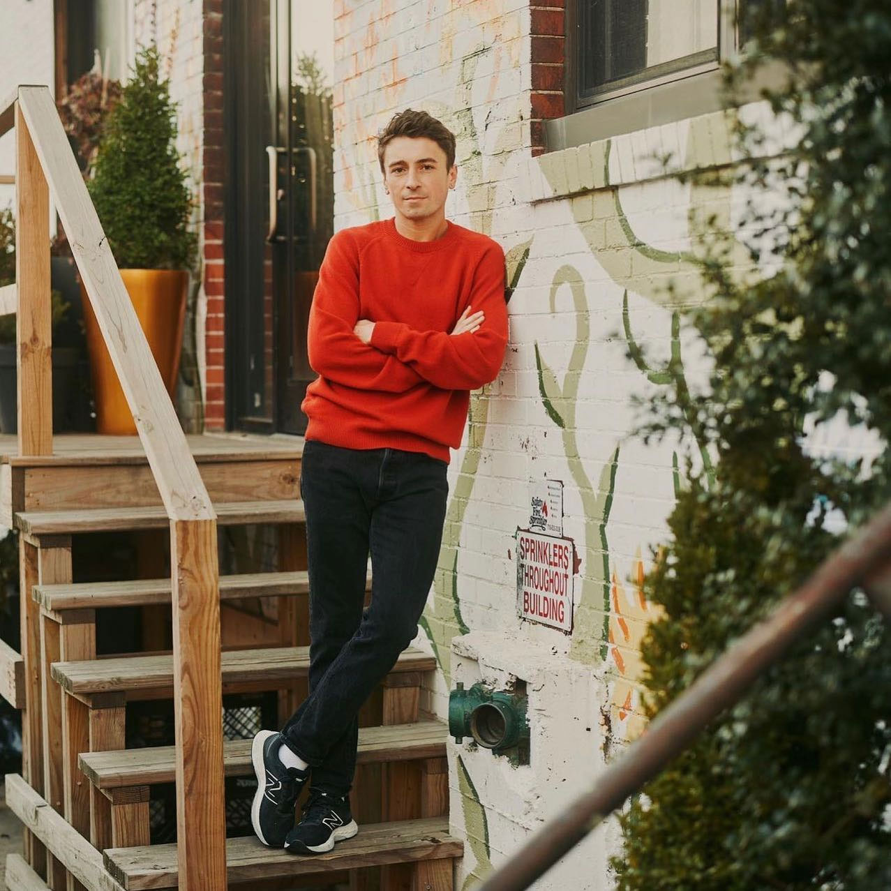 Eine Person in einem roten Pullover und dunklen Jeans lehnt an einer Wand mit bunten Graffiti. Sie steht auf Holzstufen, in deren Nähe Pflanzen stehen.