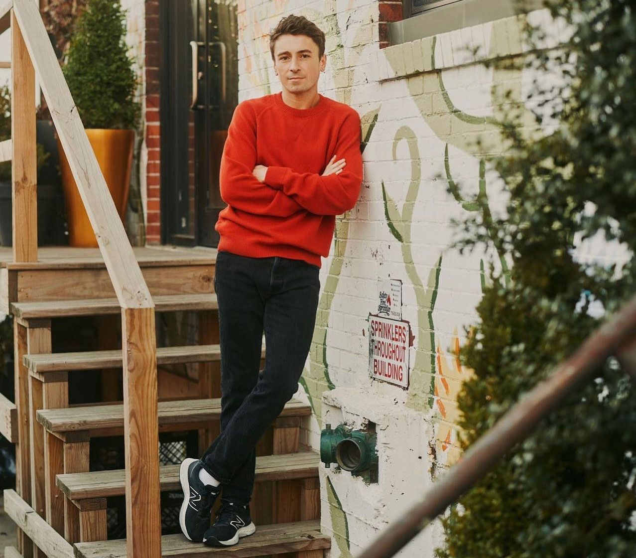 Eine Person in rotem Pullover und dunkler Hose lehnt an einer weißen Backsteinmauer mit Efeumuster und steht auf Holzstufen. Ein kleines Schild mit der Aufschrift „Sprinkleranlage im gesamten Gebäude“ ist zu sehen. Im Hintergrund sind grüne Töpfe zu sehen.