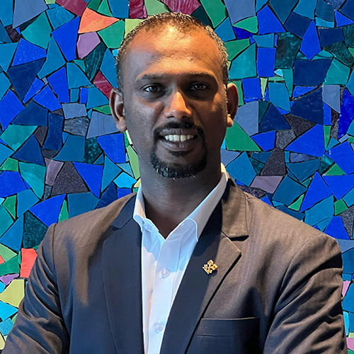 A man in a dark suit and white shirt stands smiling with arms crossed in front of a colorful mosaic wall made of various blue, green, and purple tile pieces.