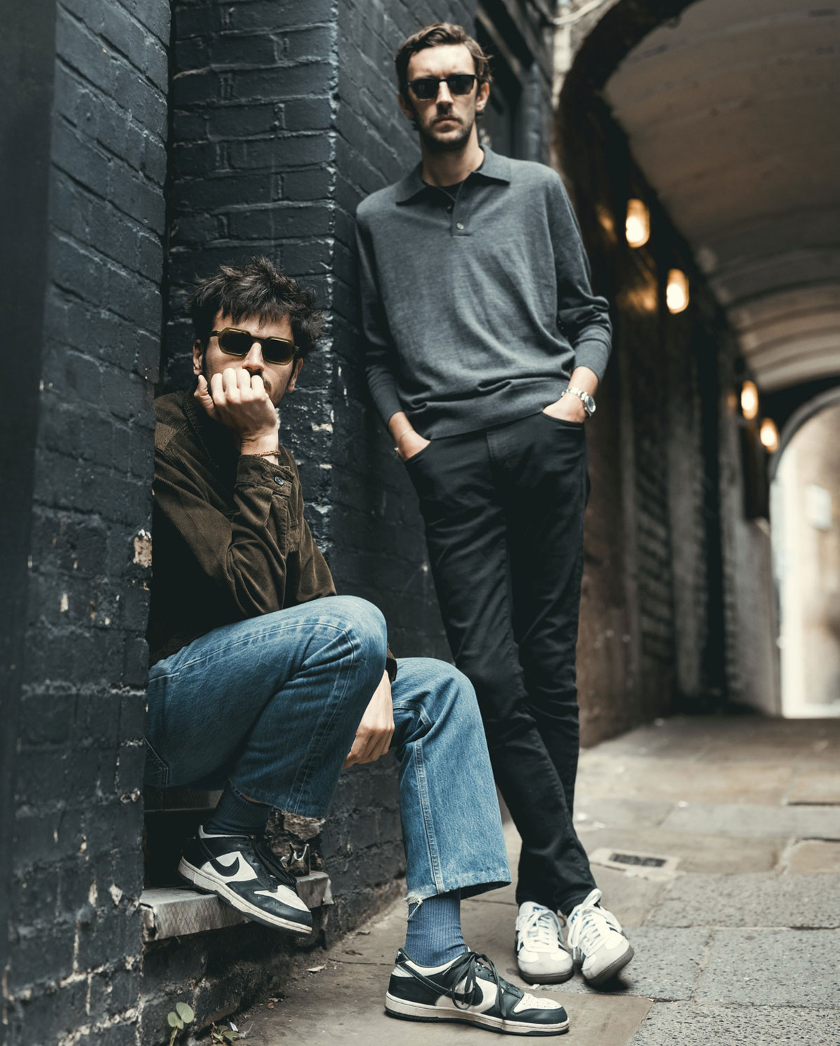 Two men wearing sunglasses pose in an urban alley; one sits against a brick wall in jeans and sneakers, while the other stands with hands in pockets, dressed in dark pants and a grey sweater. The scene is moody and stylish.