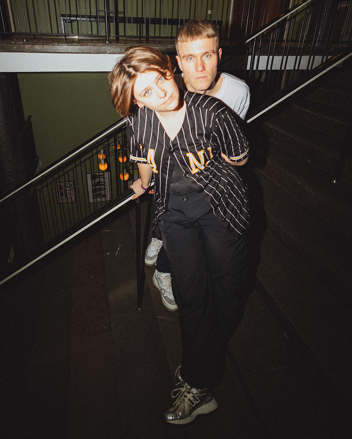 Two people pose closely together on a staircase indoors; the person in front leans back slightly while wearing a black pinstripe shirt, and the person behind wears a white T-shirt. Both look directly at the camera.