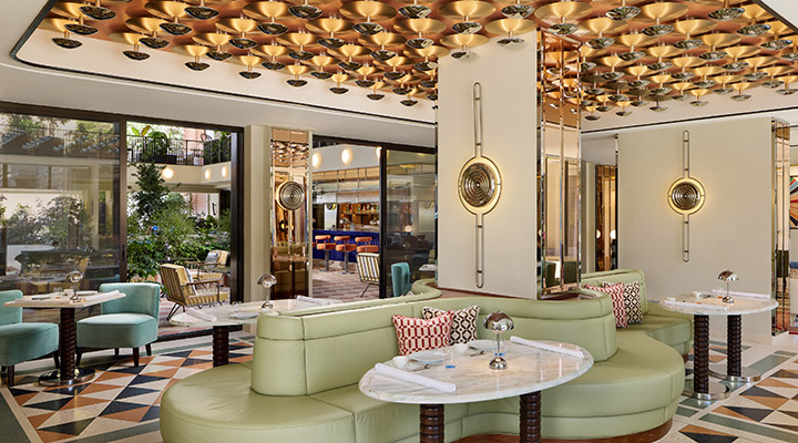 Chic hotel lounge with mint-green curved sofas, round marble tables, patterned floor, gold ceiling décor, geometric cushions, and retro-style wall lights; open area with view of bar and greenery outside.