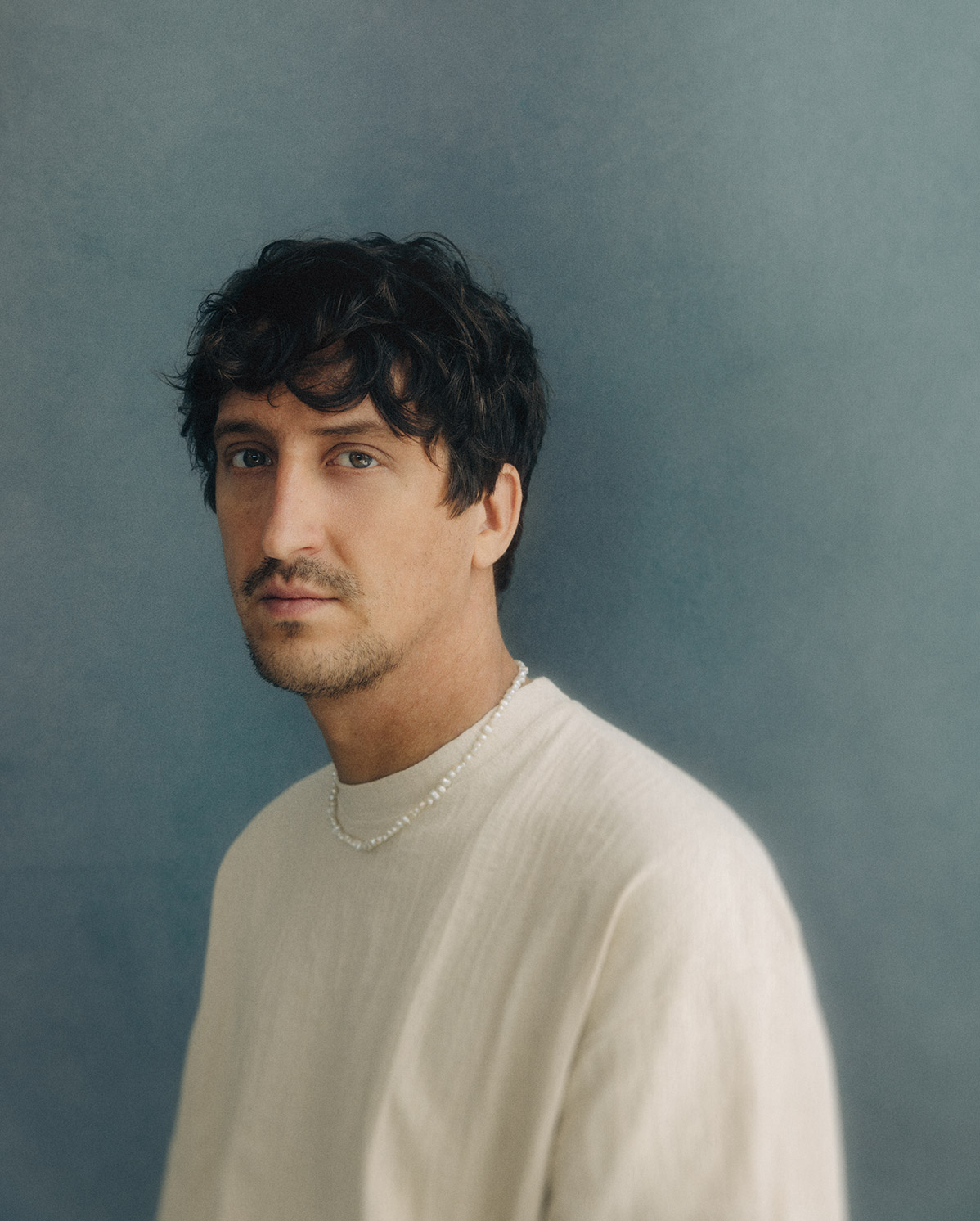 A man with wavy dark hair and light stubble, wearing a cream-colored shirt and a beaded necklace, poses in front of a soft blue background, looking slightly to the side.