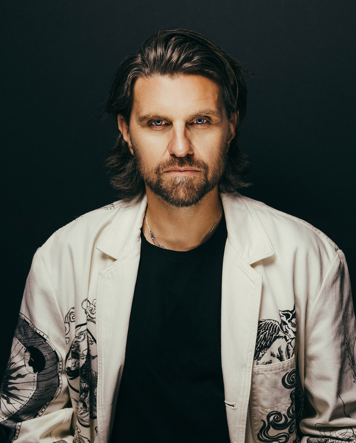 A man with shoulder-length brown hair and a beard stares seriously at the camera. He is wearing a black shirt and a light-colored jacket with dark artistic patterns on it, set against a dark background.