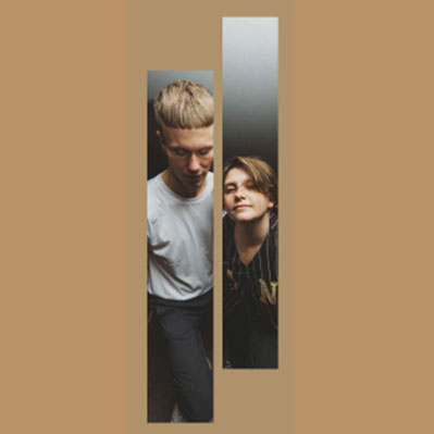 Two people are seen through two vertical rectangular cutouts against a tan background. One is standing and looking down on the left, the other is kneeling and smiling on the right.