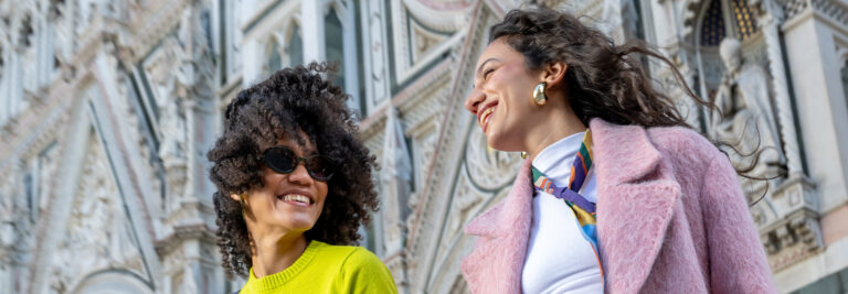 Two women smiling at each other, dressed in colorful outfits, standing in front of an ornate, historic building with intricate designs.