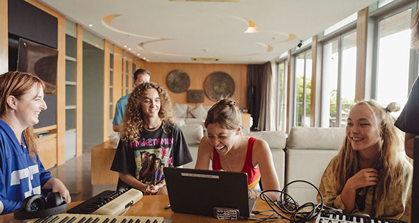 Four young women are sitting around a table with music equipment and a laptop, laughing and smiling in a bright, modern living room with large windows.